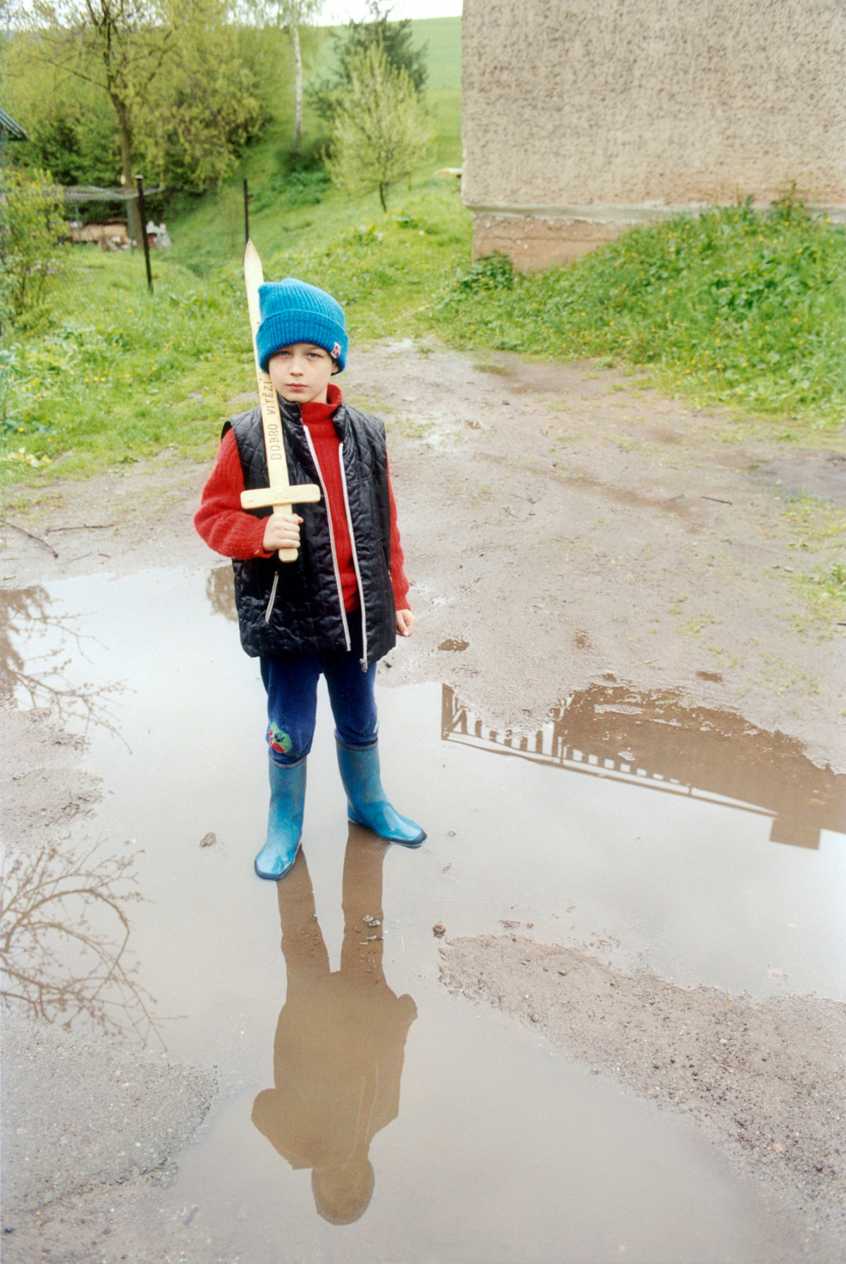 Ein kleiner Junge mit blauer Mütze und blauen Gummistiefeln steht in einer Regenpfütze und hält ein Holzschwert in der Hand. Das Schwert hat er über seine Schulter gelehnt.